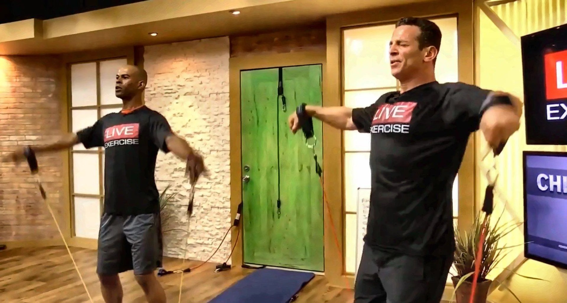 Two Bodylastics trainers lead a follow-along resistance band workout in the studio, with bands anchored to a door behind them.