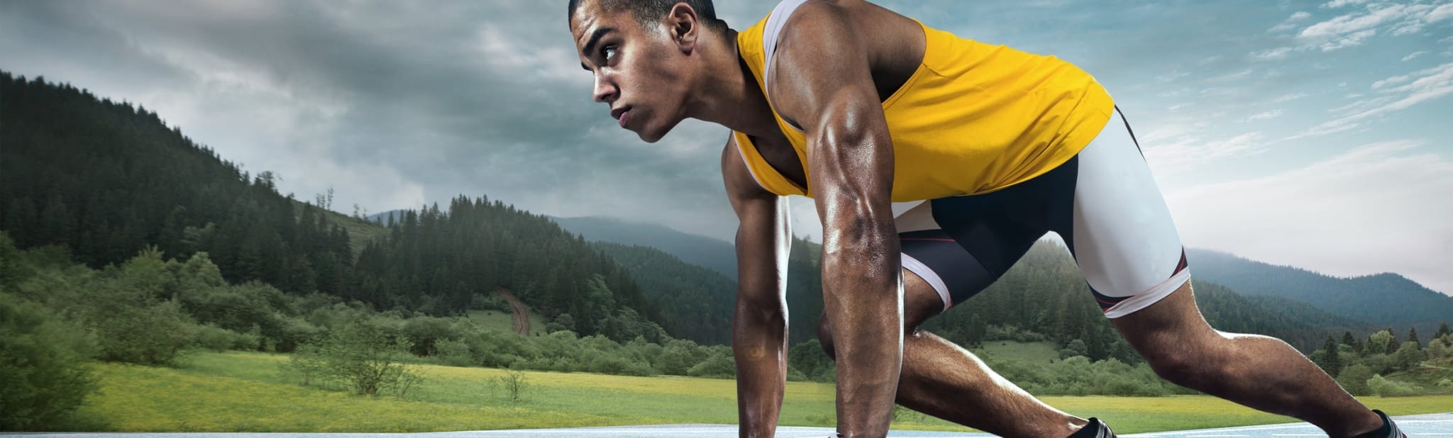 A sprinter crouches in the starting position on a track, with green hills and a cloudy sky behind him.