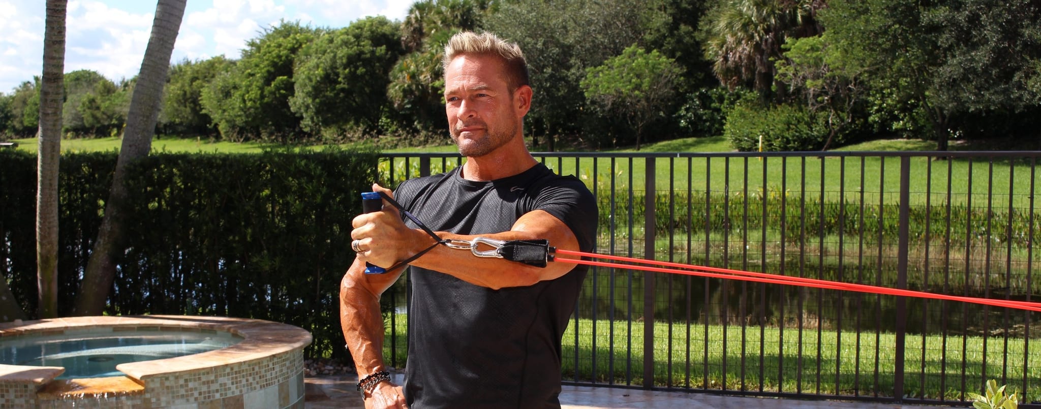 A man stands outdoors by a pool and pulls a Bodylastics band handle across his chest, with red bands stretched to an anchor behind him.